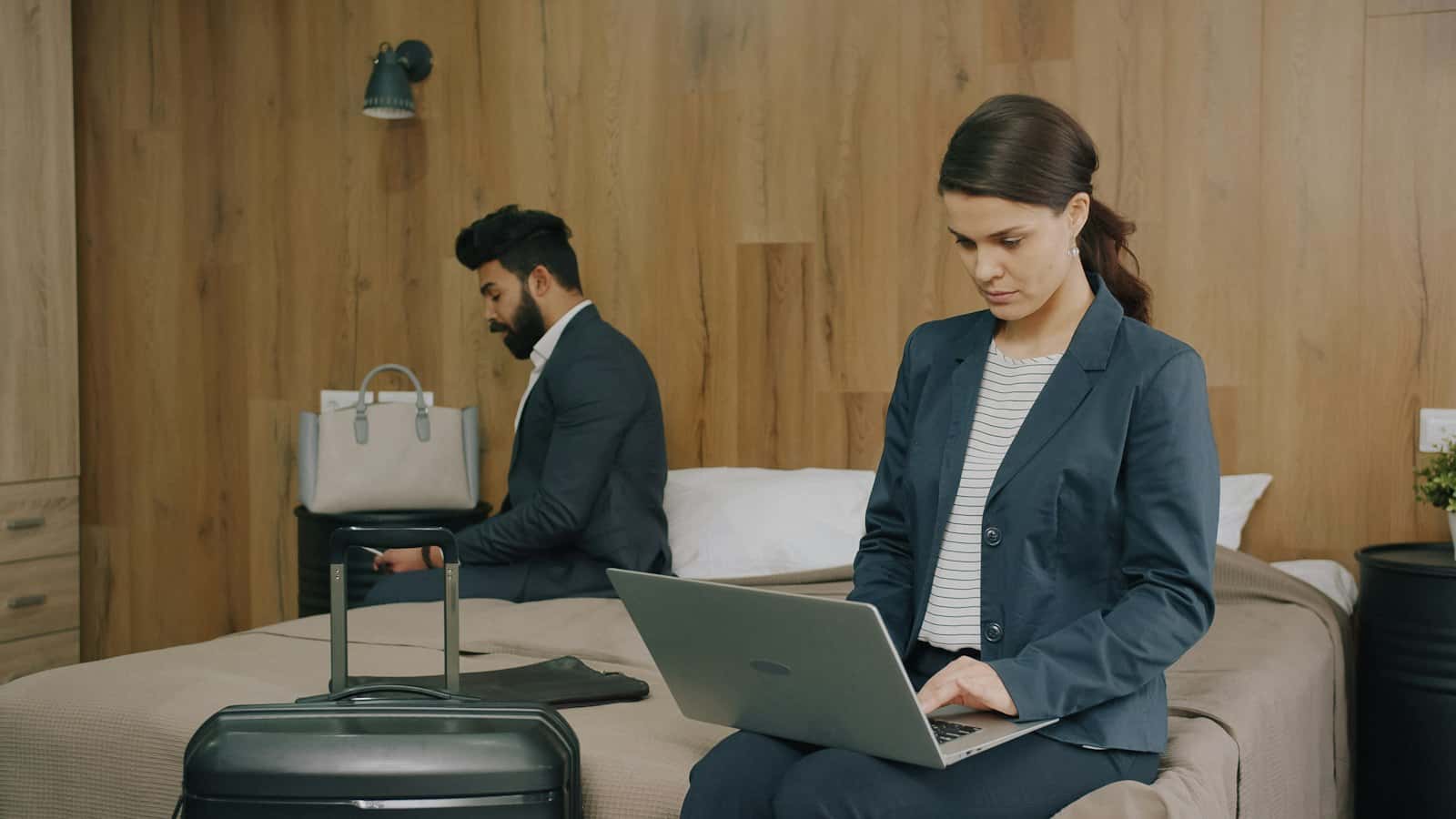 A guest on a hotel bed booking on a laptop, a suitcase beside her